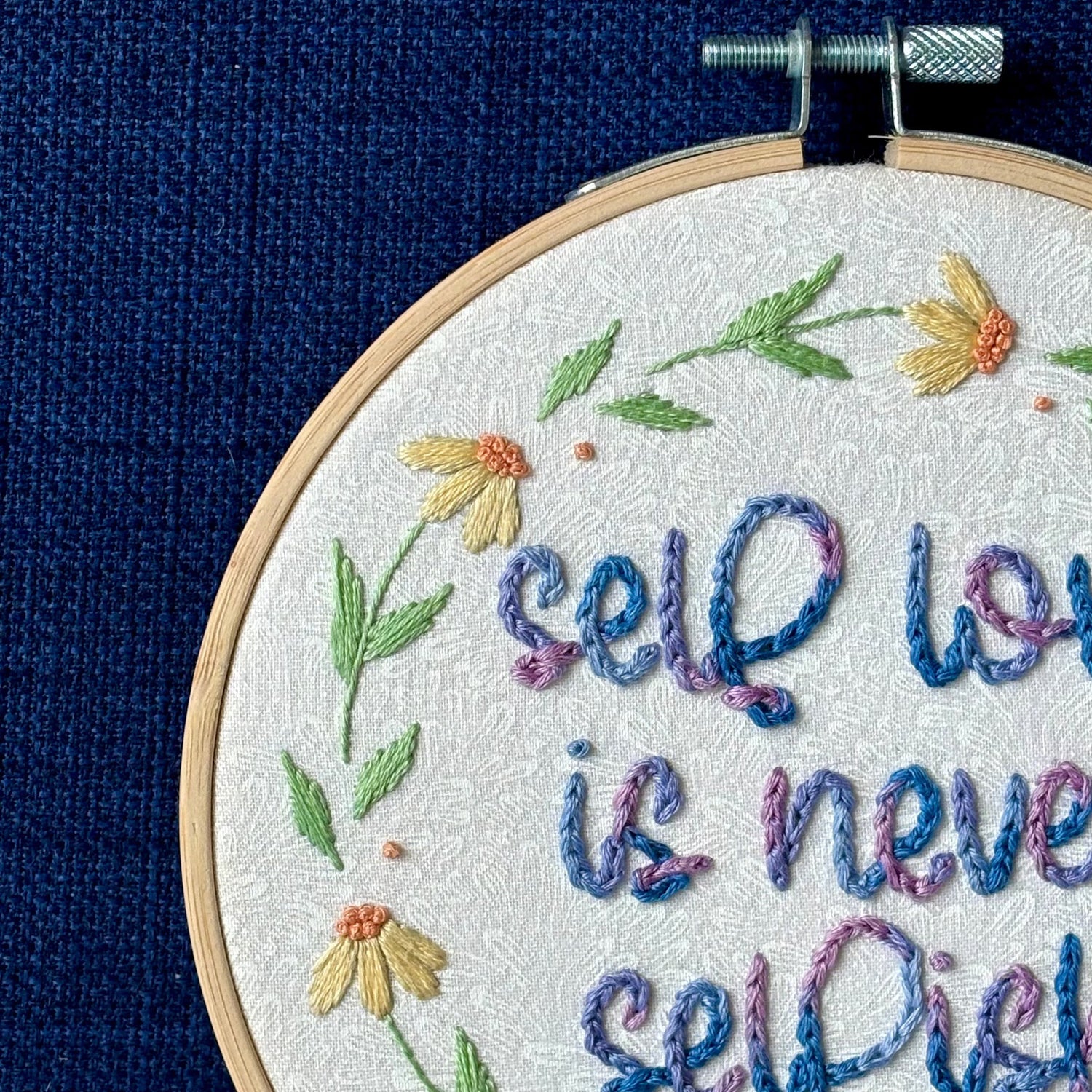 "self love is never selfish." Quote hand embroidered in color changing thread of pink, lavender, light blue, and royal blue. Surrounding the quote is a floral wreath of yellow and orange daisies. Sewn on white-on-white cotton. Finished wall art is framed in a light wood embroidery hoop.