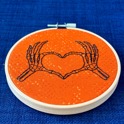 Hand embroidered pair of skeleton hands making a heart by touching thumbs and forefingers, while remaining fingers fan out. Bones are outlined with fine black embroidery. Stitched on bright orange cotton and displayed in light wooden hoop as wall art or Christmas ornament