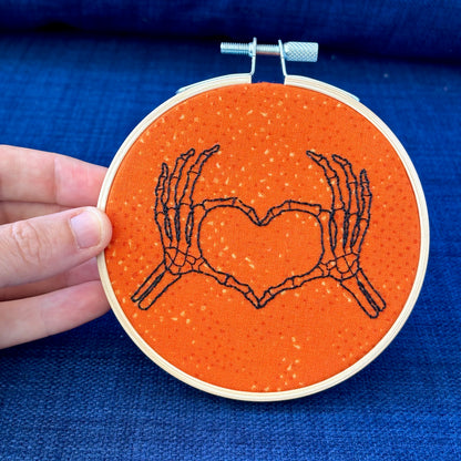 Hand embroidered pair of skeleton hands making a heart by touching thumbs and forefingers, while remaining fingers fan out. Bones are outlined with fine black embroidery. Stitched on bright orange cotton and displayed in light wooden hoop as wall art or Christmas ornament