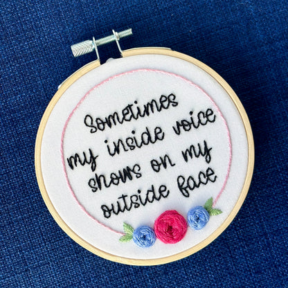 Completed embroidery hoop reads, "Sometimes my inside voice shows on my outside face". Quote in handwriting font and hand embroidered in black. Words are centered in a floral frame, which is light pink and embellished with cheerful fuchsia and periwinkle roses and light green leaves sewn with embroidery thread. Fabric is white cotton. Framed in light wood embroidery hoop and displayed as wall art or Christmas ornament.