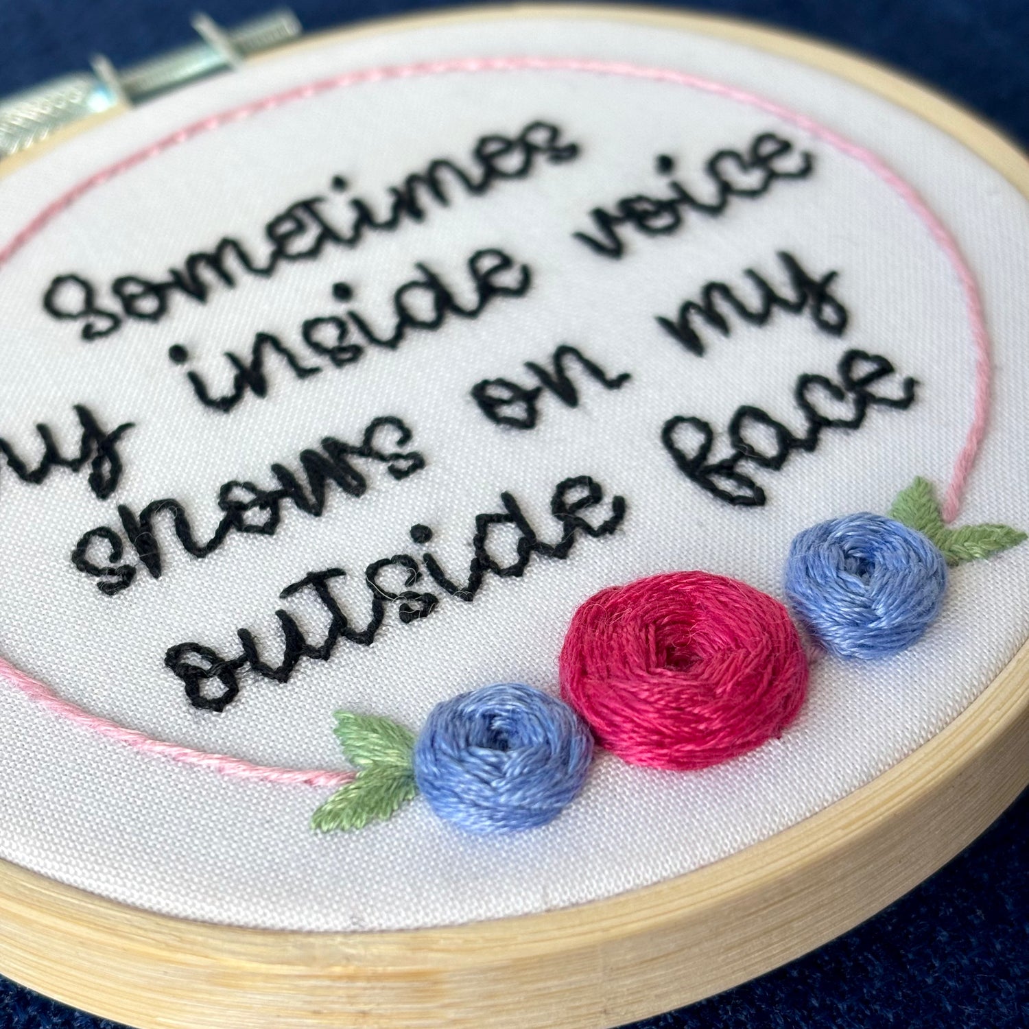 Completed embroidery hoop reads, "Sometimes my inside voice shows on my outside face". Quote in handwriting font and hand embroidered in black. Words are centered in a floral frame, which is light pink and embellished with cheerful fuchsia and periwinkle roses and light green leaves sewn with embroidery thread. Fabric is white cotton. Framed in light wood embroidery hoop and displayed as wall art or Christmas ornament.