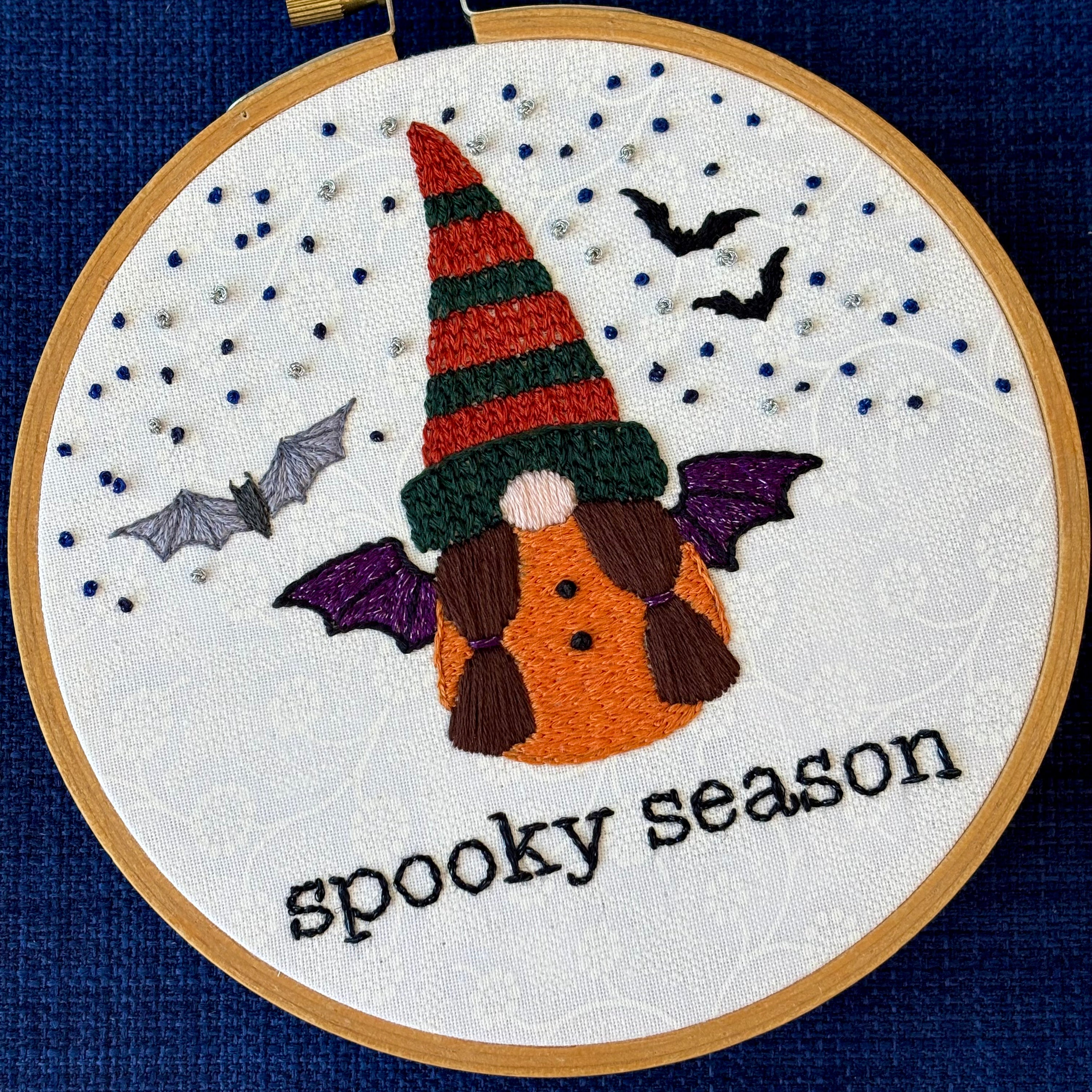 Hand embroidered gnome with dark green and burnt orange knit hat. She has brown hair in two ponytails and a pink nose. She wears a an orange robe with two black buttons and black and sparkly purple bat wings. In the background are sparkle blue and metallic silver stars, the silhouettes of two bats, and a closer detailed gray bat. Under the gnome is "spooky season" embroidered in sparkling black thread. Displayed in light wooden hoop on white cotton as wall art