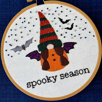 Hand embroidered gnome with dark green and burnt orange knit hat. She has brown hair in two ponytails and a pink nose. She wears a an orange robe with two black buttons and black and sparkly purple bat wings. In the background are sparkle blue and metallic silver stars, the silhouettes of two bats, and a closer detailed gray bat. Under the gnome is "spooky season" embroidered in sparkling black thread. Displayed in light wooden hoop on white cotton as wall art