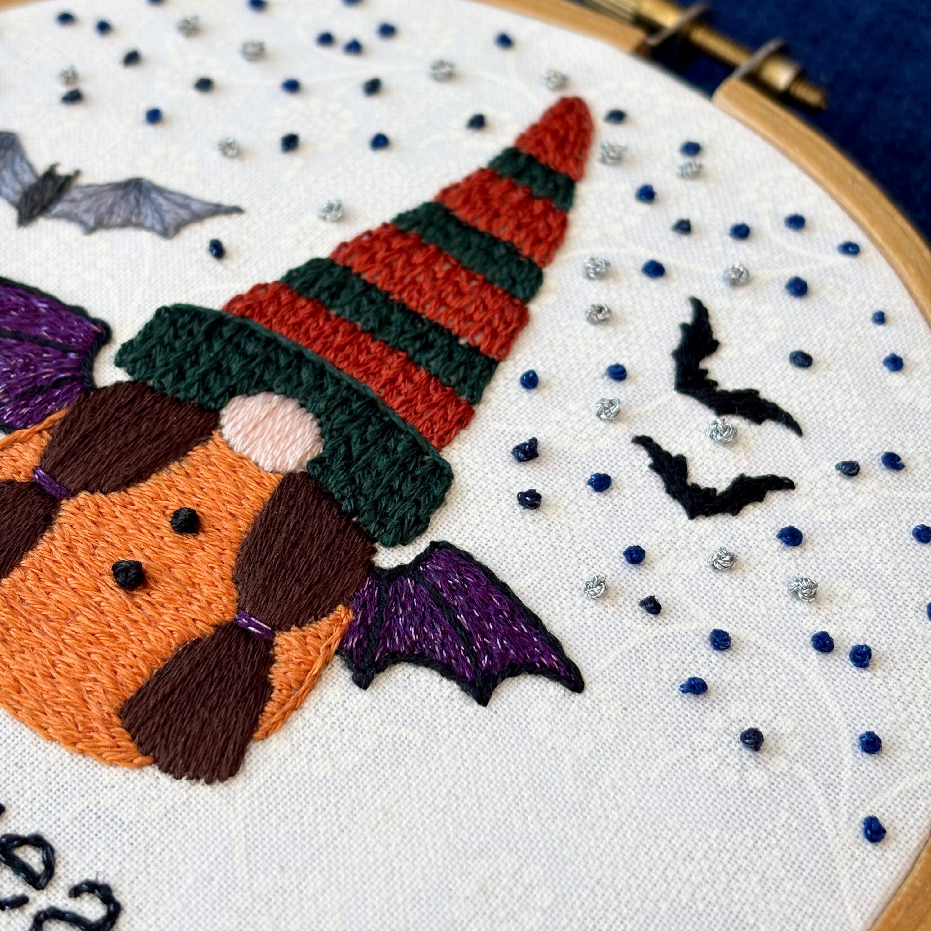 Hand embroidered gnome with dark green and burnt orange knit hat. She has brown hair in two ponytails and a pink nose. She wears a an orange robe with two black buttons and black and sparkly purple bat wings. In the background are sparkle blue and metallic silver stars, the silhouettes of two bats, and a closer detailed gray bat. Under the gnome is "spooky season" embroidered in sparkling black thread. Displayed in light wooden hoop on white cotton as wall art