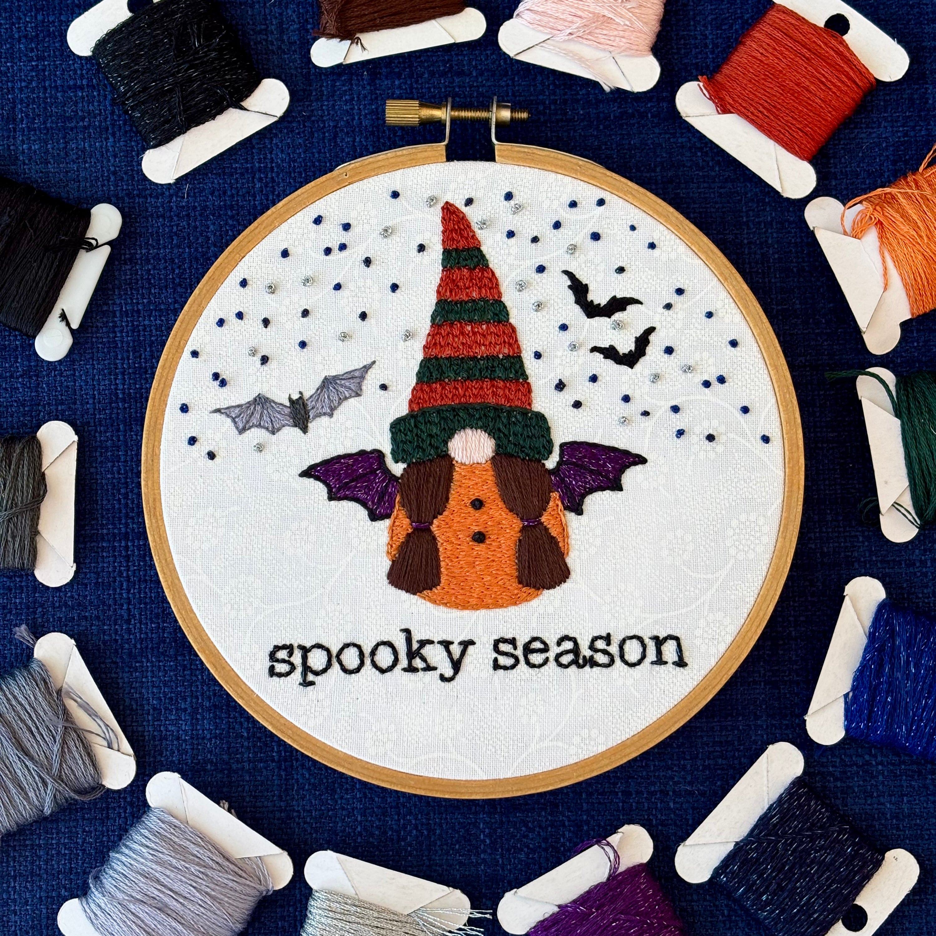 Hand embroidered gnome with dark green and burnt orange knit hat. She has brown hair in two ponytails and a pink nose. She wears a an orange robe with two black buttons and black and sparkly purple bat wings. In the background are sparkle blue and metallic silver stars, the silhouettes of two bats, and a closer detailed gray bat. Under the gnome is "spooky season" embroidered in sparkling black thread. Displayed in light wooden hoop on white cotton as wall art