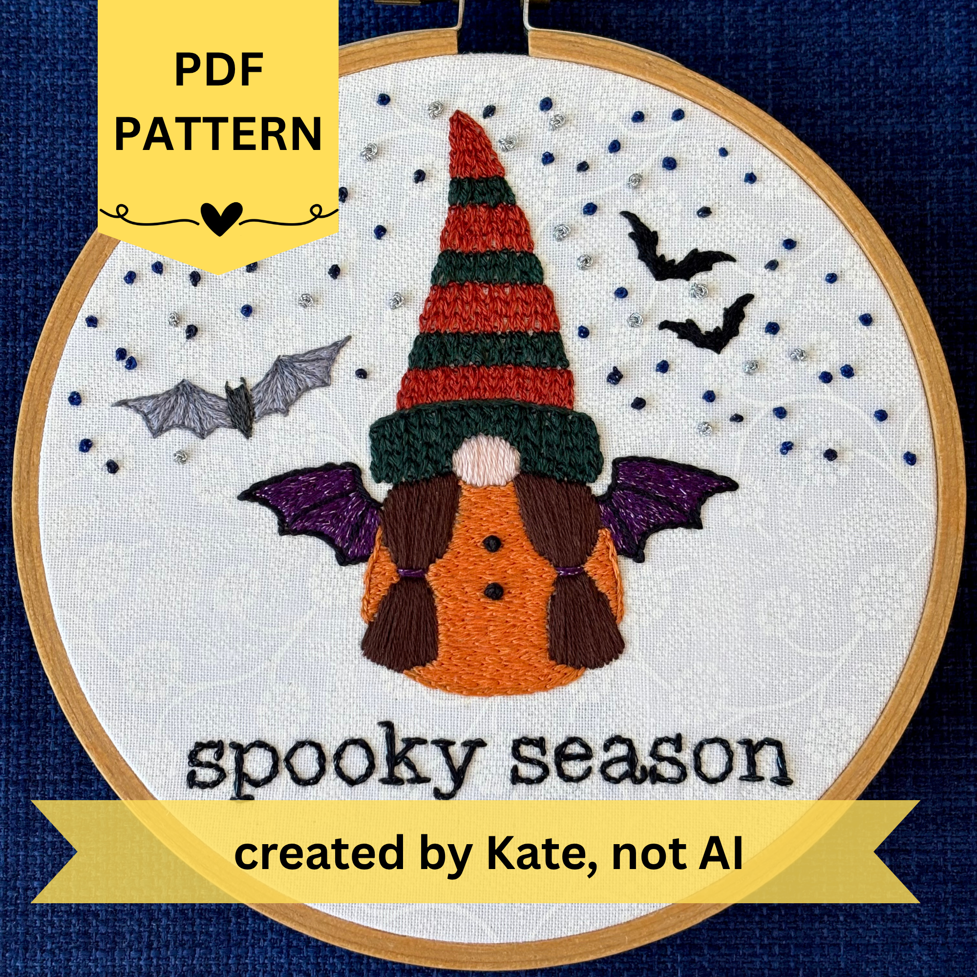 PDF pattern created by Kate not AI. Hand embroidered gnome with dark green and burnt orange knit hat. She has brown hair in two ponytails and a pink nose. She wears a an orange robe with two black buttons and black and sparkly purple bat wings. In the background are sparkle blue and metallic silver stars, the silhouettes of two bats, and a closer detailed gray bat. Under the gnome is "spooky season" embroidered in sparkling black thread. Displayed in light wooden hoop on white cotton as wall art