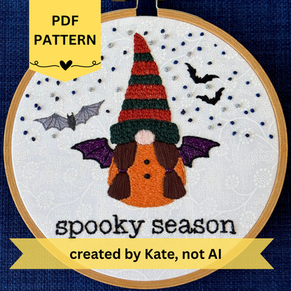 PDF pattern created by Kate not AI. Hand embroidered gnome with dark green and burnt orange knit hat. She has brown hair in two ponytails and a pink nose. She wears a an orange robe with two black buttons and black and sparkly purple bat wings. In the background are sparkle blue and metallic silver stars, the silhouettes of two bats, and a closer detailed gray bat. Under the gnome is "spooky season" embroidered in sparkling black thread. Displayed in light wooden hoop on white cotton as wall art