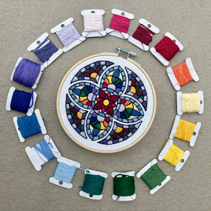 Many colored hand embroidery based on traditional stained glass windows with flowers and rainbows in the design. Fabric is white on white cotton. Embroidery is displayed in light wood embroidery hoop as wall art and surrounded by 19 DMC embroidery thread bobbins of all the colors used in the design.