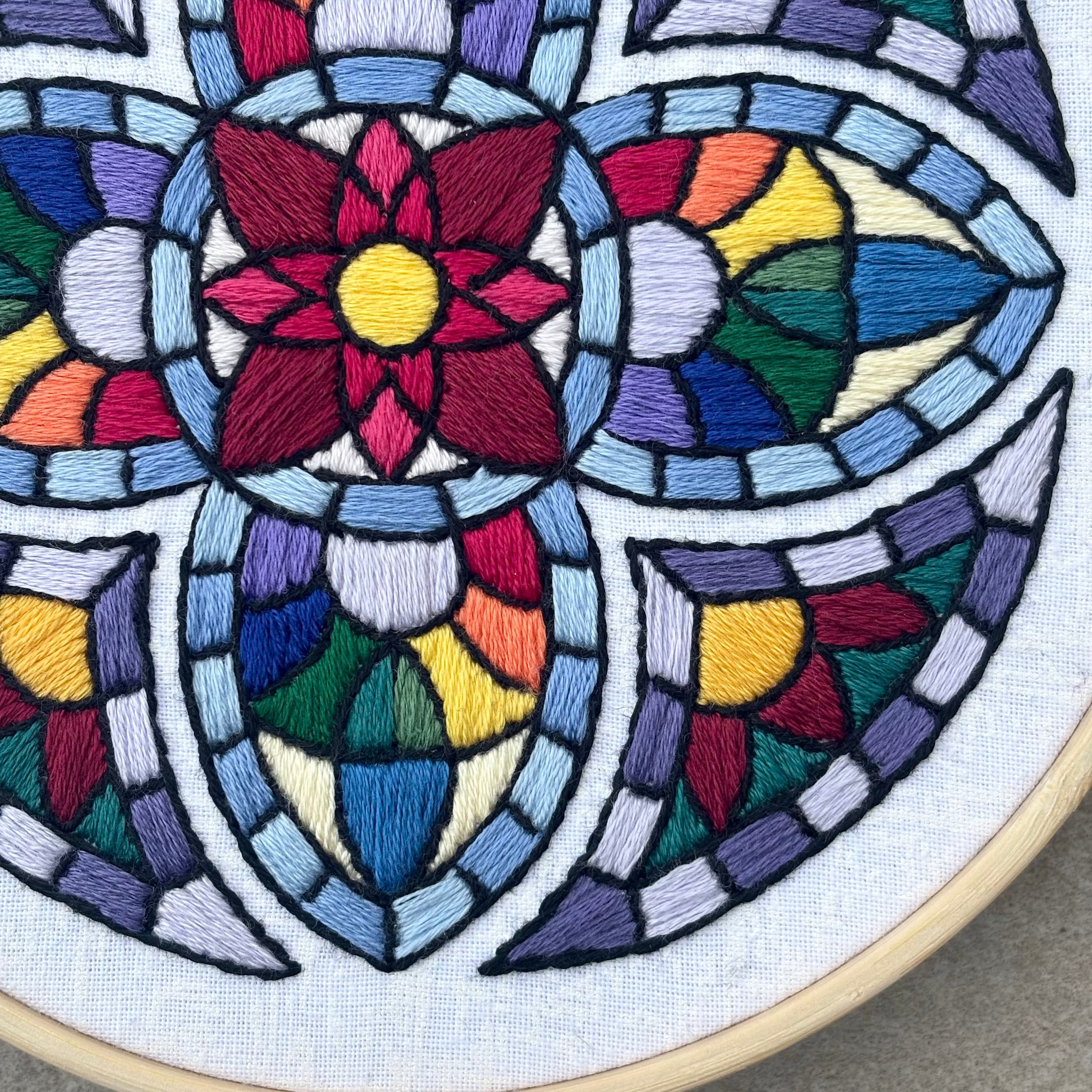 PDF Pattern created by Kate, not AI. Many colored hand embroidery based on traditional stained glass windows with flowers and rainbows in the design. Fabric is white on white cotton. Embroidery is displayed in light wood embroidery hoop as wall art.
