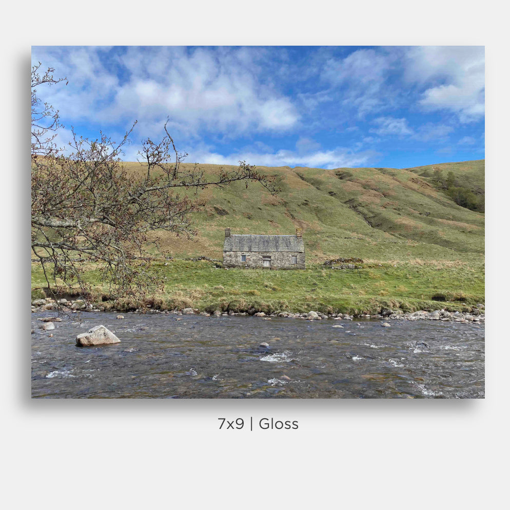 This striking landscape photograph captures a charming stone cottage on the banks of the River Tilt in Scotland, viewed from across the river beneath a brilliant blue Highland sky. The weathered cottage, though worn by time, retains a quiet beauty and character that reflects the rich history and enduring spirit of the Scottish countryside.