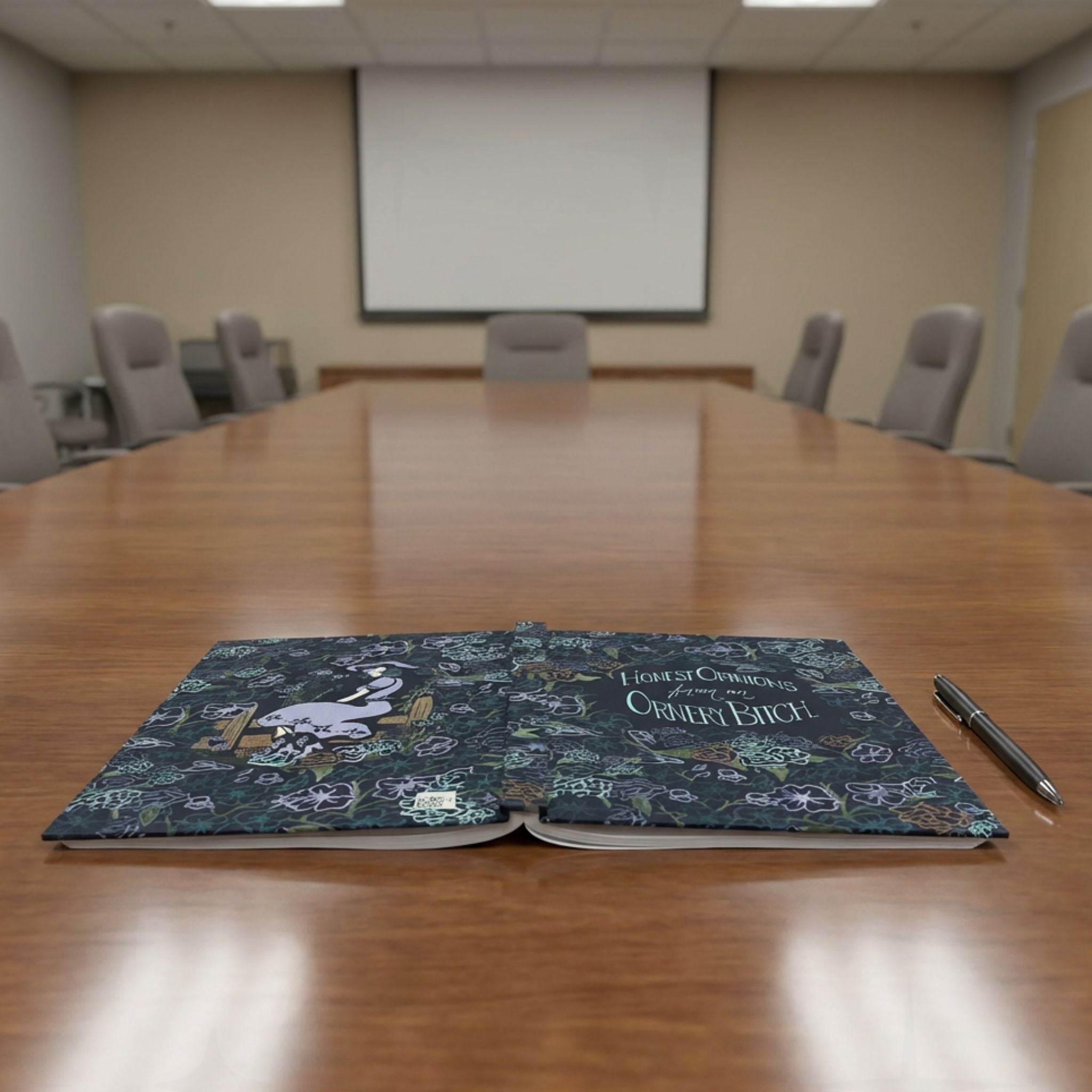 Ornery Bitch sweary journal open flat on conference room table beside a pen, showing full wrap dark floral and cowgirl pinup artwork on both covers