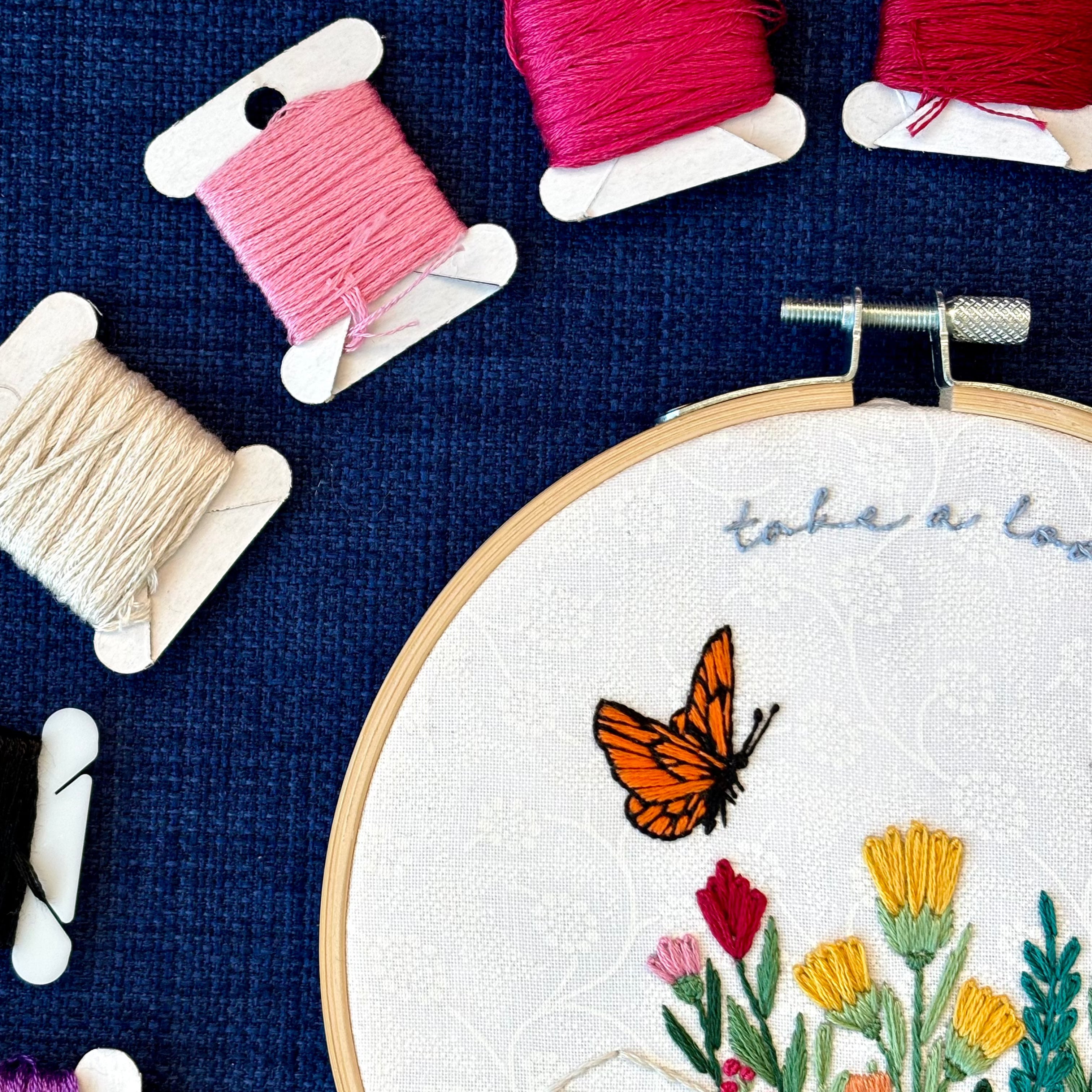 Photo of a hand embroidered design inspired by Reading Rainbow. Fabric is white cotton with white flowers printed on it. There is an open book with a rainbow of flowers growing from it. Hovering above are an orange and black butterfly and a bright blue and black butterfly. The top right curve of the pattern reads, "take a look, it&