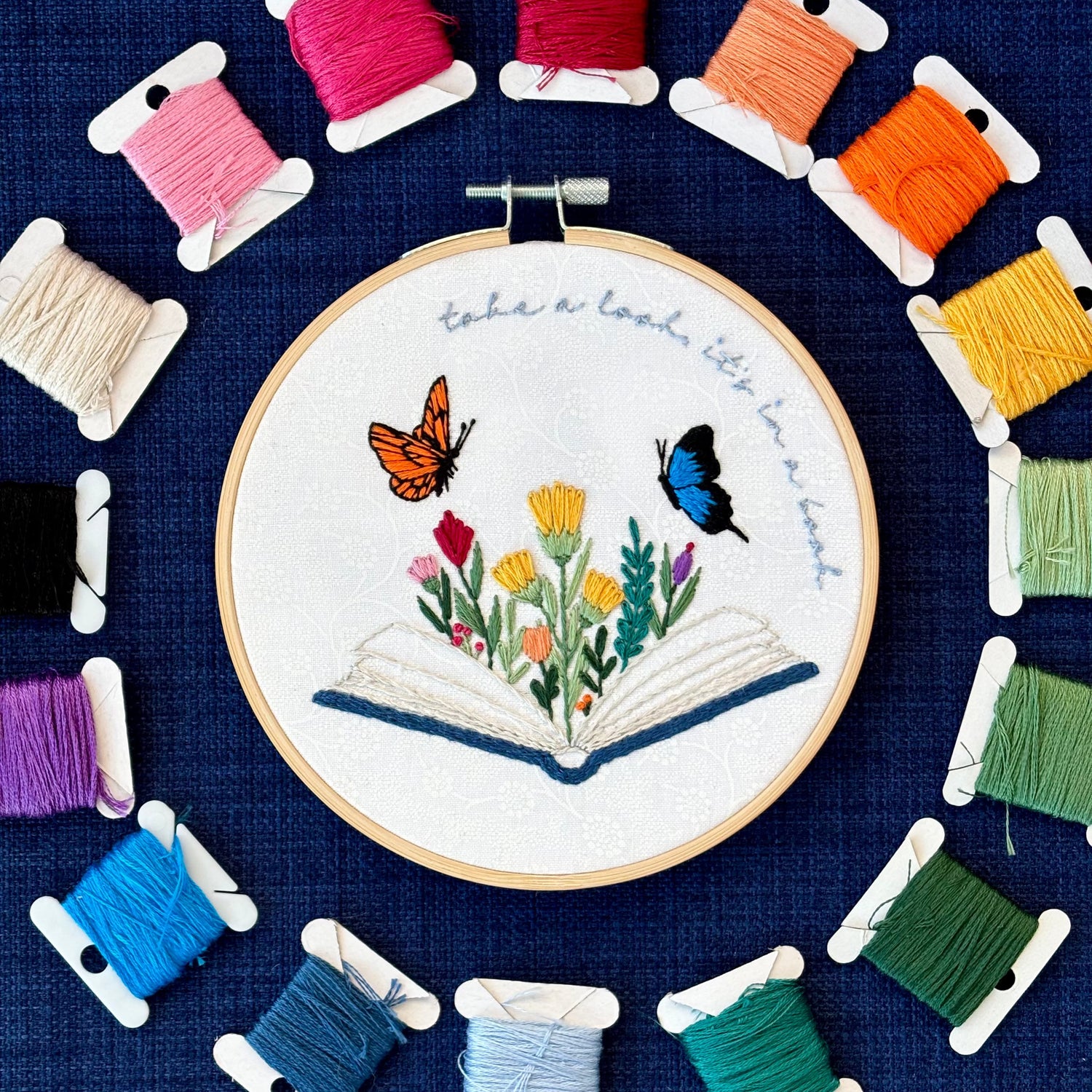 Photo of a hand embroidered design inspired by Reading Rainbow. Fabric is white cotton with white flowers printed on it. There is an open book with a rainbow of flowers growing from it. Hovering above are an orange and black butterfly and a bright blue and black butterfly. The top right curve of the pattern reads, "take a look, it&