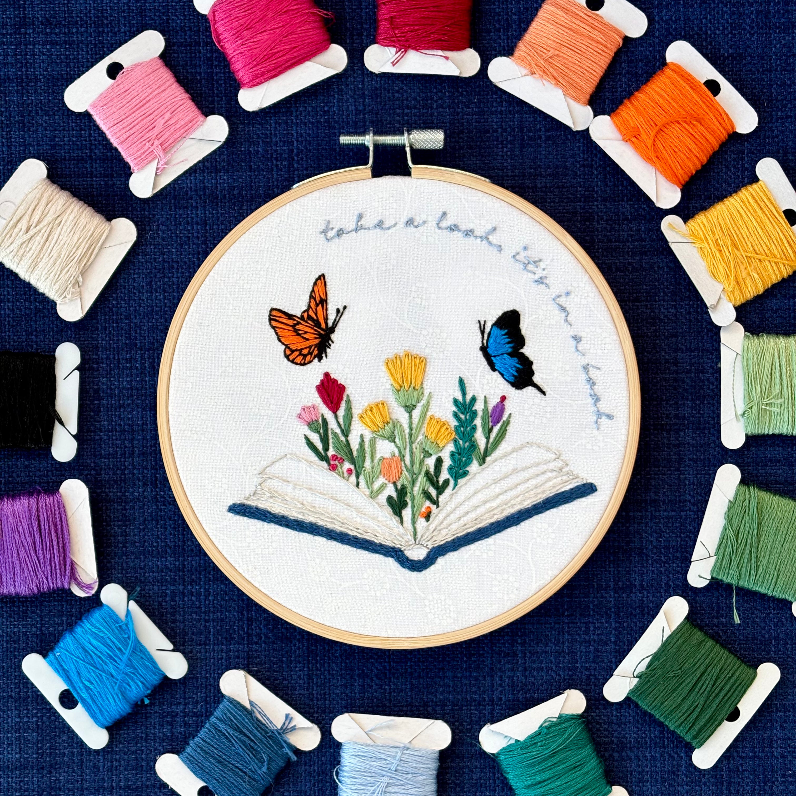 Photo of a hand embroidered design inspired by Reading Rainbow. Fabric is white cotton with white flowers printed on it. There is an open book with a rainbow of flowers growing from it. Hovering above are an orange and black butterfly and a bright blue and black butterfly. The top right curve of the pattern reads, "take a look, it&
