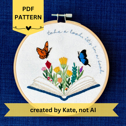 PDF pattern created by Kate, not AI. Photo of a hand embroidered design inspired by Reading Rainbow. Fabric is white cotton with white flowers printed on it. There is an open book with a rainbow of flowers growing from it. Hovering above are an orange and black butterfly and a bright blue and black butterfly. The top right curve of the pattern reads, "take a look, it&