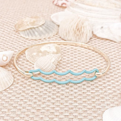 the beach and back double aqua wave tension bangle in gold tone on neutral color woven textured table top with white seashells scattered in the background