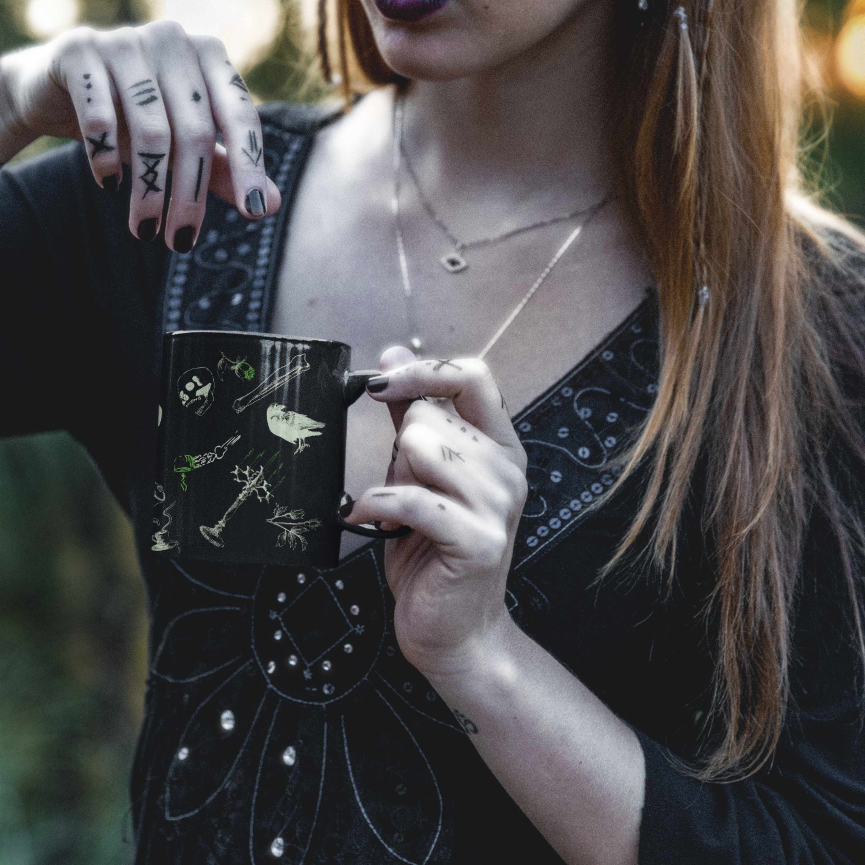 Woman with tattooed hands in a black embroidered top holding a toxic green goth mug outdoors in warm light