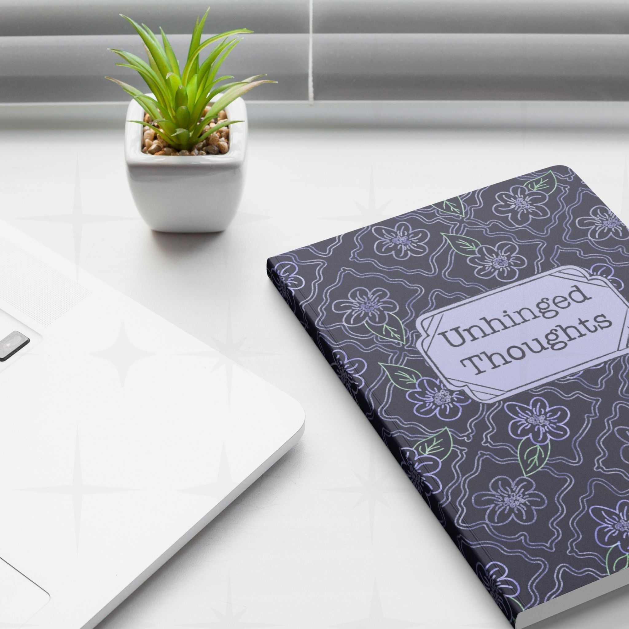 Unhinged Thoughts dark floral softcover journal placed on a desk next to a laptop and a plant