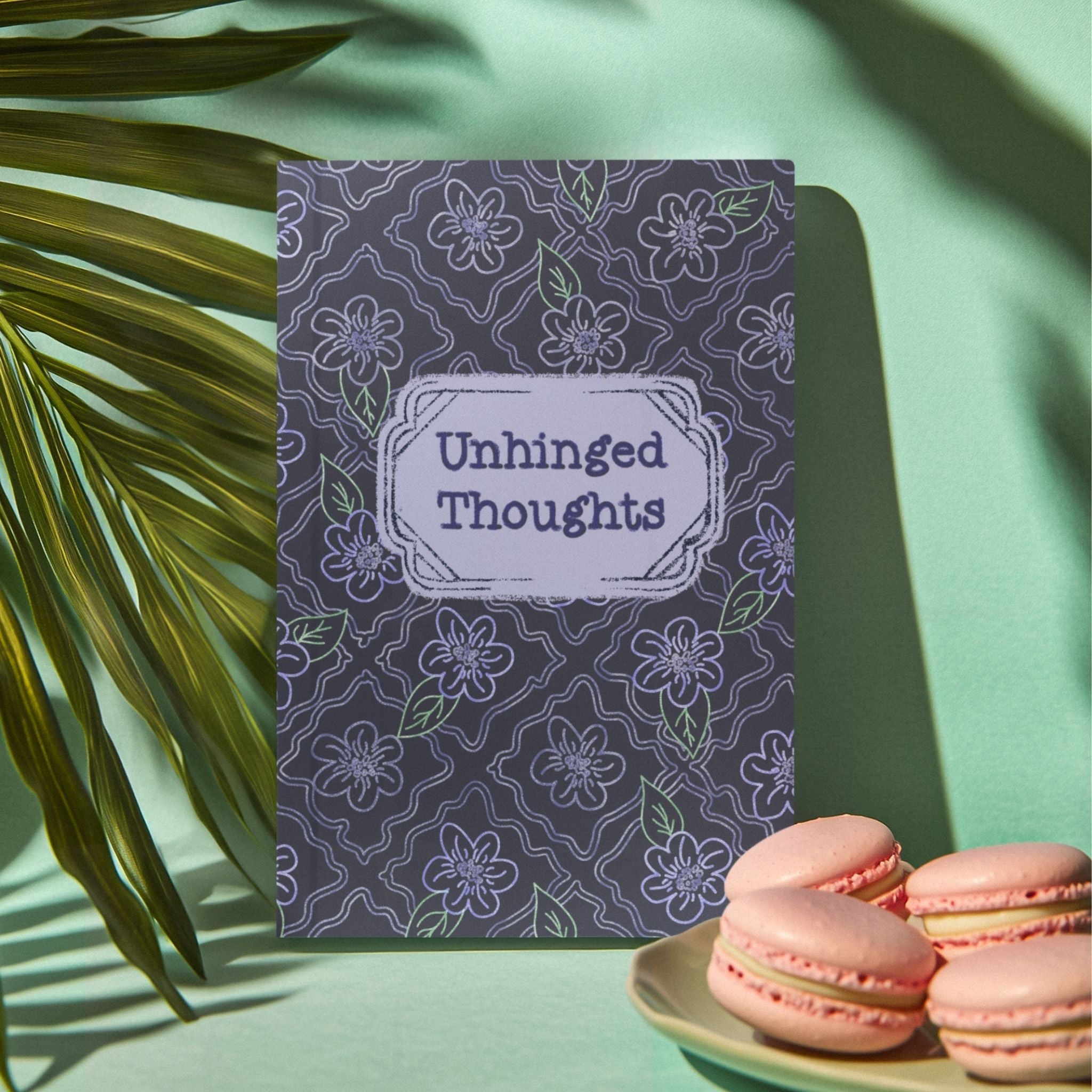 Unhinged Thoughts dark floral softcover journal standing upright beside pink macarons and palm frond on mint green background