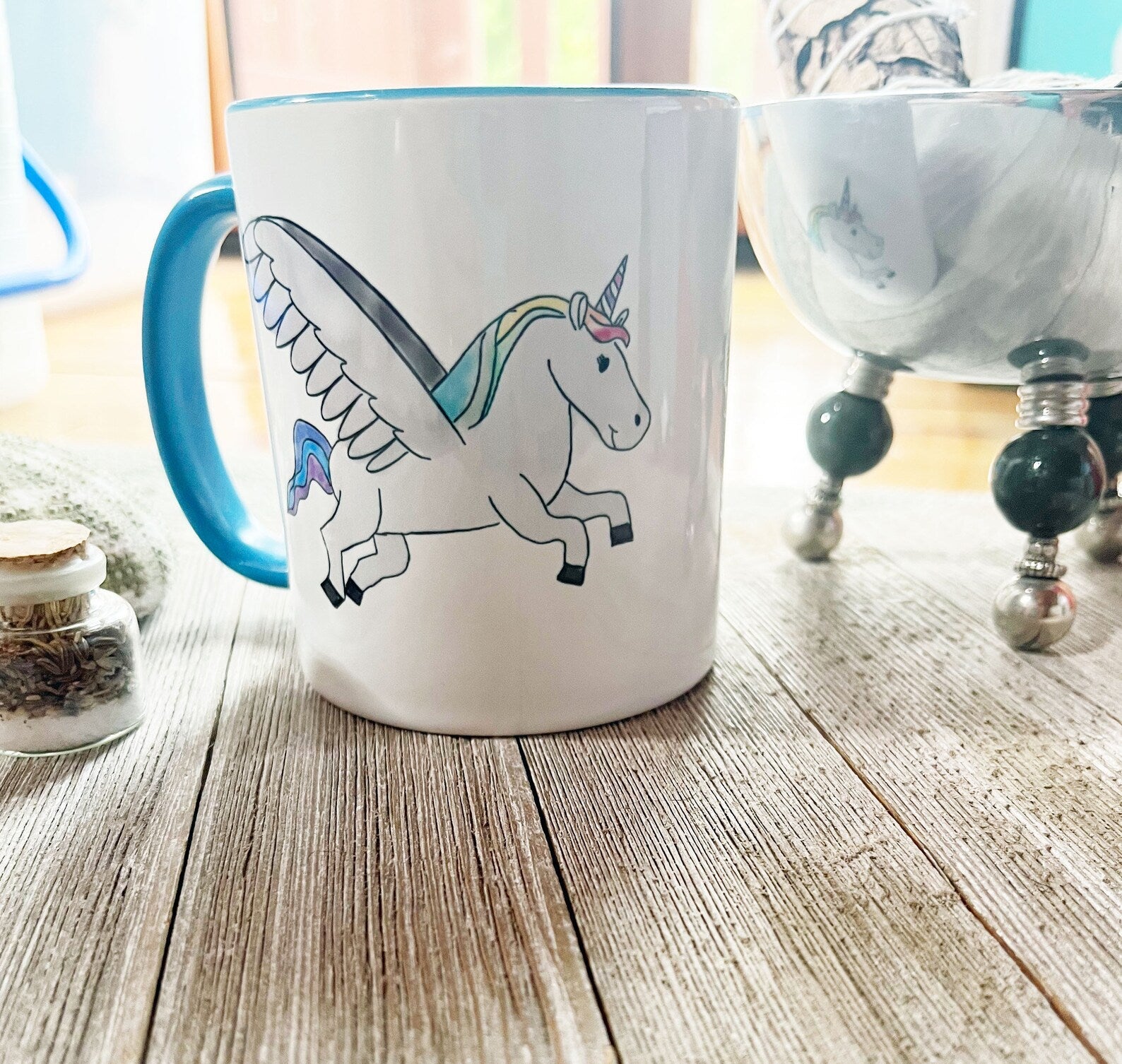The “Unicorn with Rainbow Wings Mug with Color Inside” by Enlivenadorn by Gwynstone is a vibrant ceramic mug featuring a whimsical illustration of a winged unicorn with a rainbow mane and blue handle. Resting on a wooden surface beside a small jar and reflective bowl, this fantasy animal mug makes the perfect gift for any unicorn enthusiast.