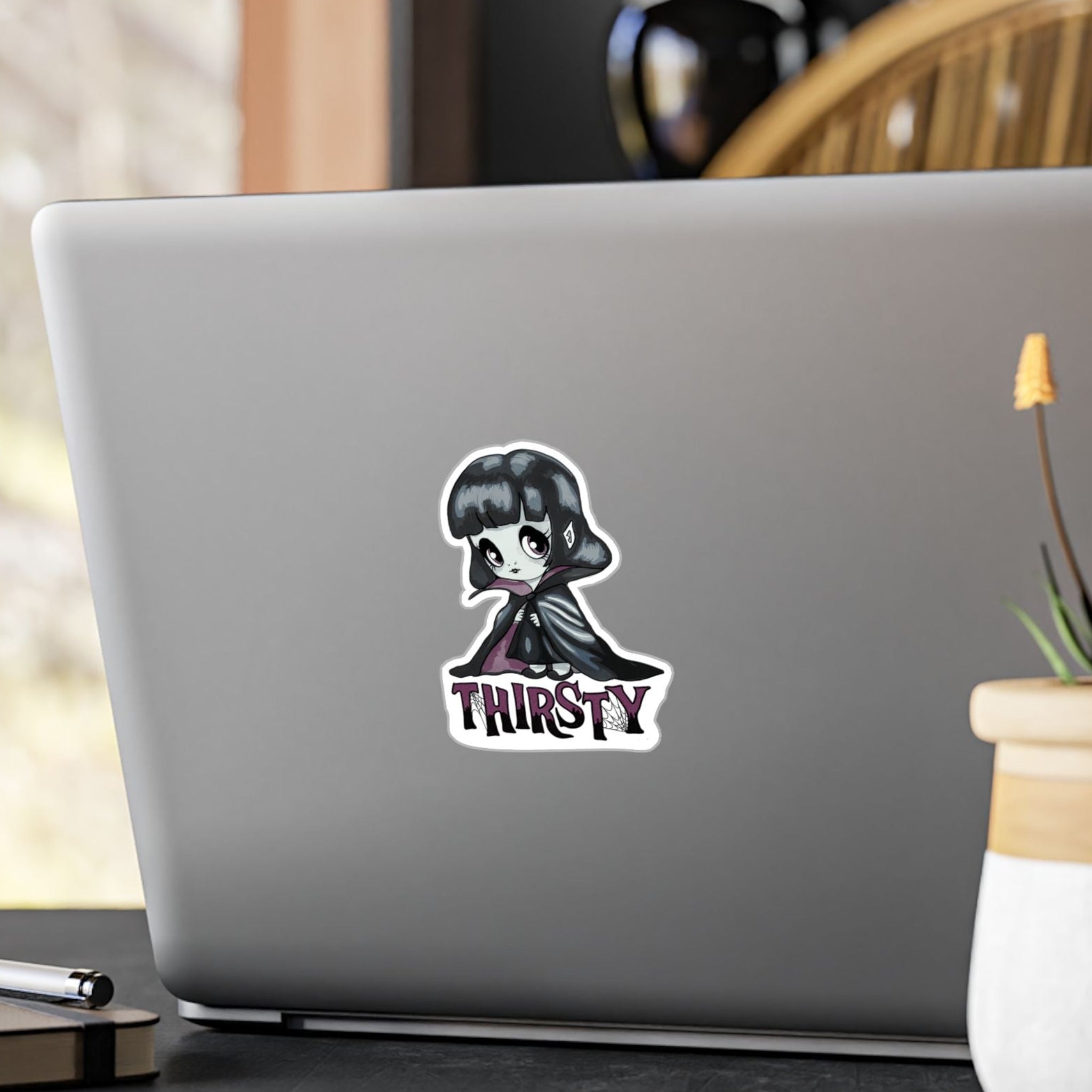 Small chibi vampire girl sticker with plum eyes and THIRSTY lettering on the back of a silver laptop