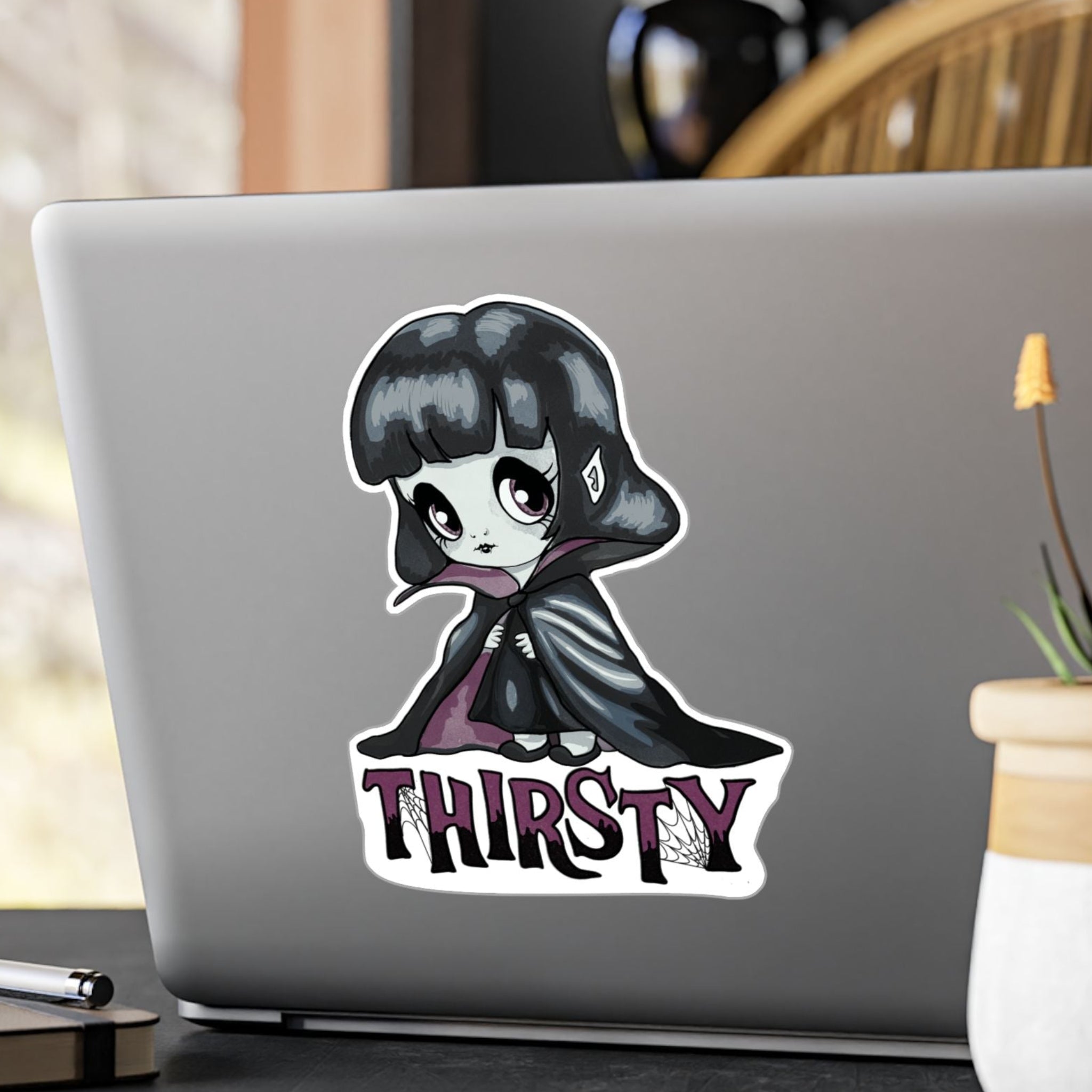 Large chibi vampire girl sticker with plum eyes and THIRSTY lettering on the back of a silver laptop