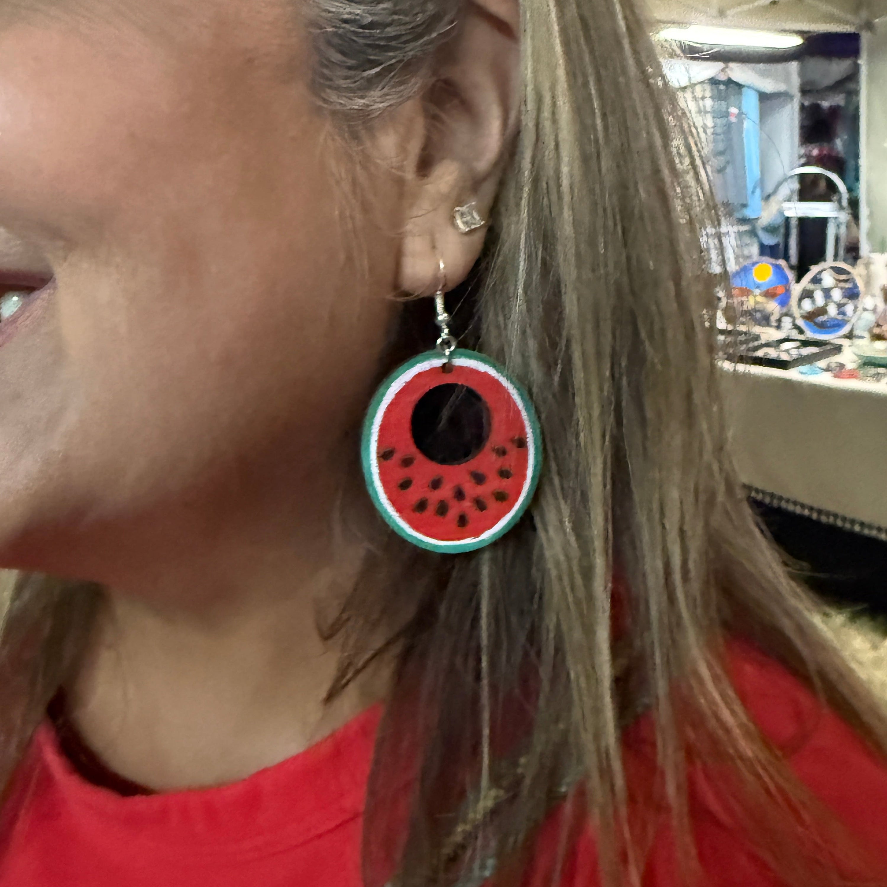 Hand-painted wooden watermelon slice dangle earring worn on the ear, featuring a vibrant red watermelon design with black seeds, a white rind, and a green outer edge. This close-up showcases the lightweight construction and hypoallergenic .925 sterling silver hook, making it a fun summer accessory for fruit lovers, farmers markets, vacations, and everyday wear.