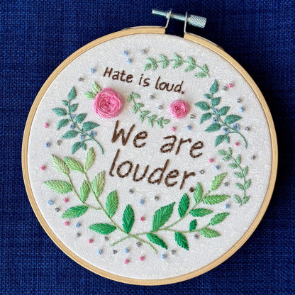 "Hate is loud. We are louder." Pride quote hand embroidered in chocolate brown. Surrounded by green leaves &amp; vines, with pink roses, blue berries, &amp; metallic silver dots. Sewn on white-on-white cotton. Framed in light wood embroidery hoop as wall art.