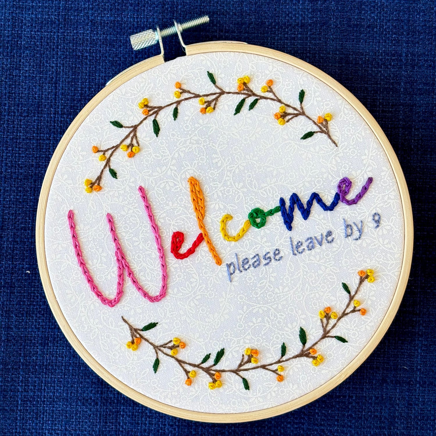 "Welcome, please leave by 9". Welcome hand embroidered in rainbow colors from pink to purple. Please leave by 9 hand embroidered in a light blue gray. A floral wreath of flower stems with yellow and orange buds create the impression of a circle. Fabric is white-on-white cotton. Framed in a light wooden embroidery hoop and displayed as wall art.