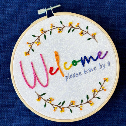 "Welcome, please leave by 9". Welcome hand embroidered in rainbow colors from pink to purple. Please leave by 9 hand embroidered in a light blue gray. A floral wreath of flower stems with yellow and orange buds create the impression of a circle. Fabric is white-on-white cotton. Framed in a light wooden embroidery hoop and displayed as wall art.