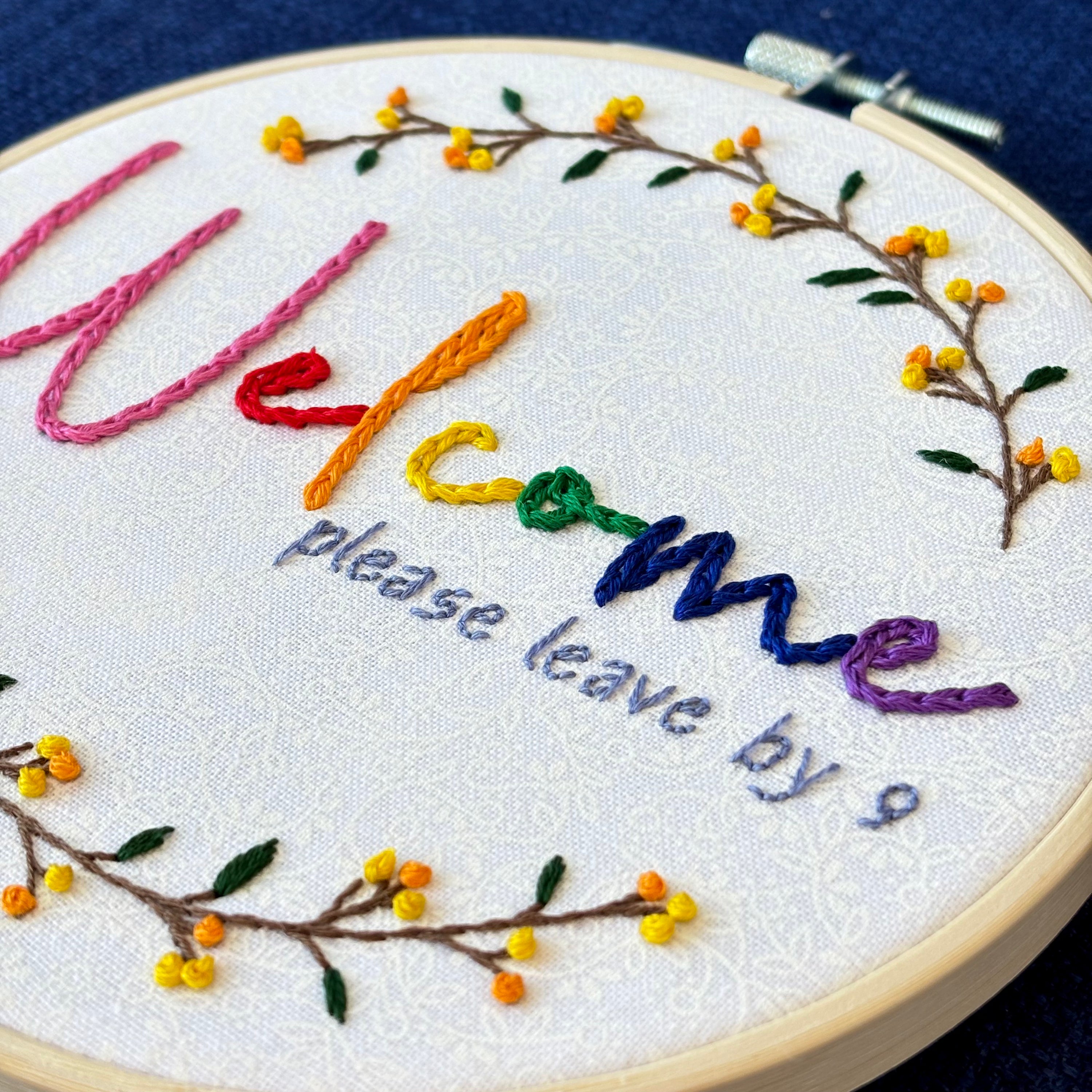 "Welcome, please leave by 9". Welcome hand embroidered in rainbow colors from pink to purple. Please leave by 9 hand embroidered in a light blue gray. A floral wreath of flower stems with yellow and orange buds create the impression of a circle. Fabric is white-on-white cotton. Framed in a light wooden embroidery hoop and displayed as wall art.