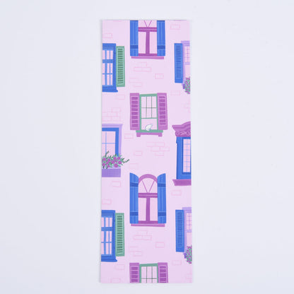A bookmark featuring a pattern of whimsical city scenes with windows, flower boxes, and a cat on a pink brick backdrop.