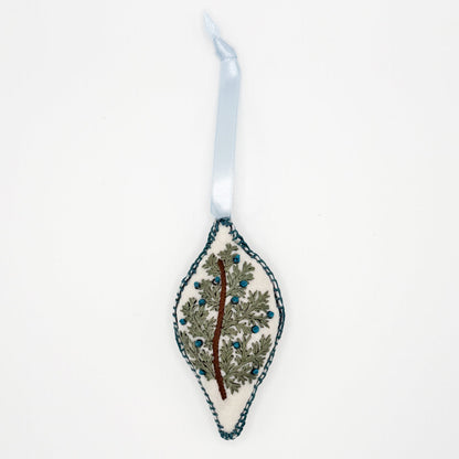 Hand embroidered and sewn oblong Christmas ornament with a brown branch, greenery and blue berries, ethically made by syrian refugee women, lay flat