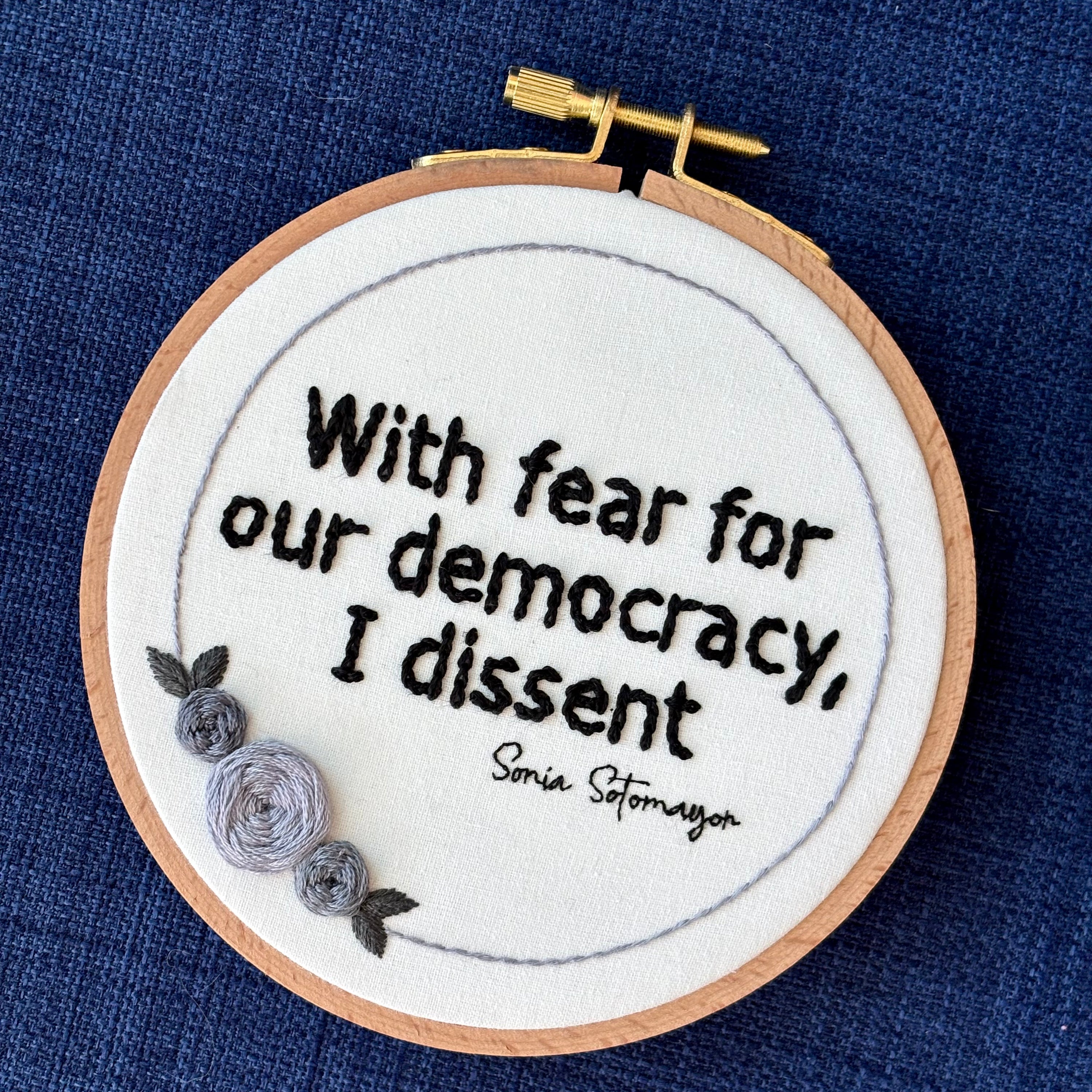 PDF Pattern created by Kate, not AI. "With fear for our democracy, I dissent. Sonia Sotomayor" Quote hand embroidered in a strong sans-serif font in black embroidery thread. A floral wreath of light and medium gray roses surrounds the text. Fabric is white cotton. Embroidery is displayed as wall art in a wood embroidery hoop.
