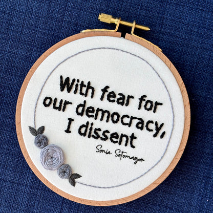 PDF Pattern created by Kate, not AI. "With fear for our democracy, I dissent. Sonia Sotomayor" Quote hand embroidered in a strong sans-serif font in black embroidery thread. A floral wreath of light and medium gray roses surrounds the text. Fabric is white cotton. Embroidery is displayed as wall art in a wood embroidery hoop.