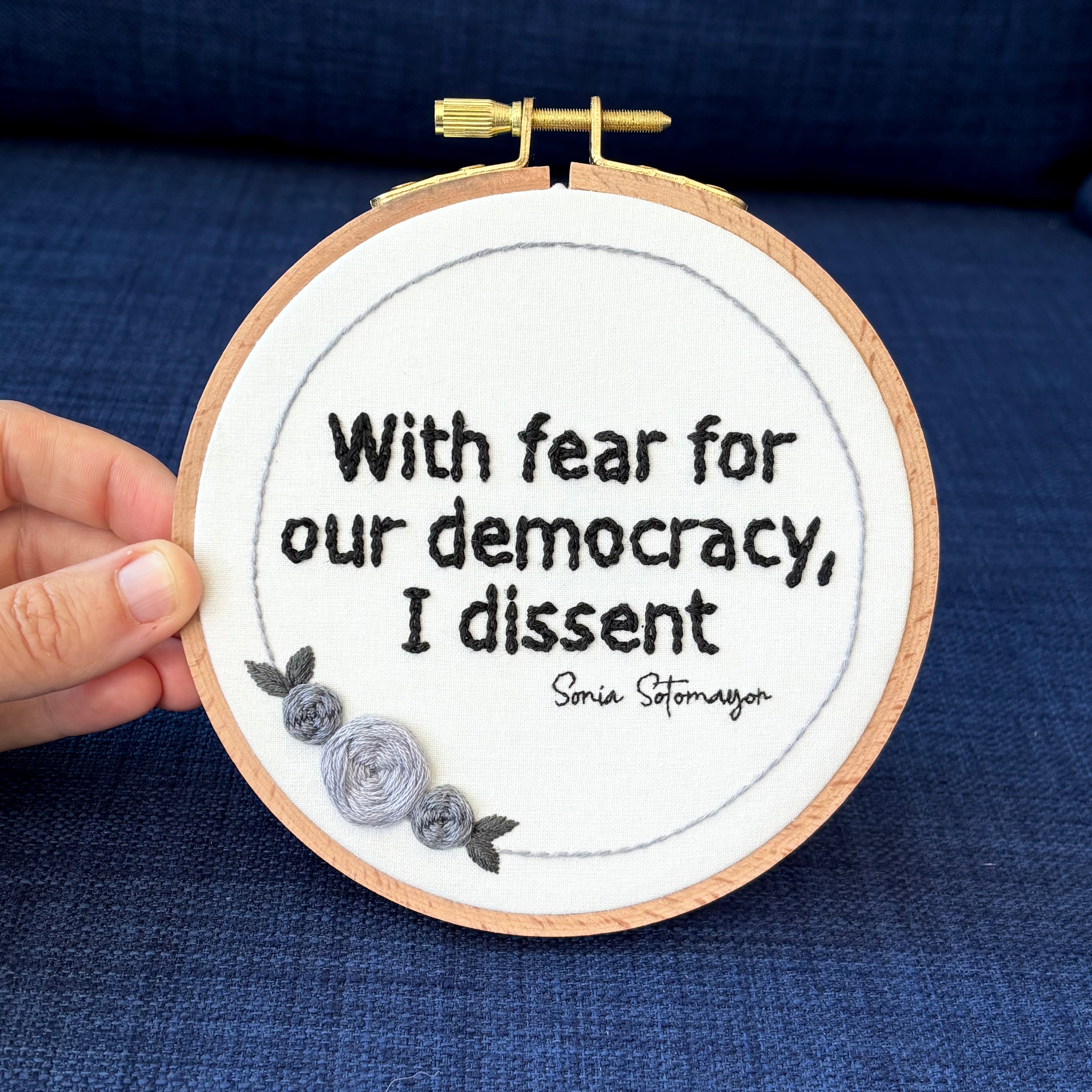 "With fear for our democracy, I dissent. Sonia Sotomayor" Quote hand embroidered in a strong sans-serif font in black embroidery thread. A floral wreath of light and medium gray roses surrounds the text. Fabric is white cotton. Embroidery is displayed as wall art in a wood embroidery hoop.