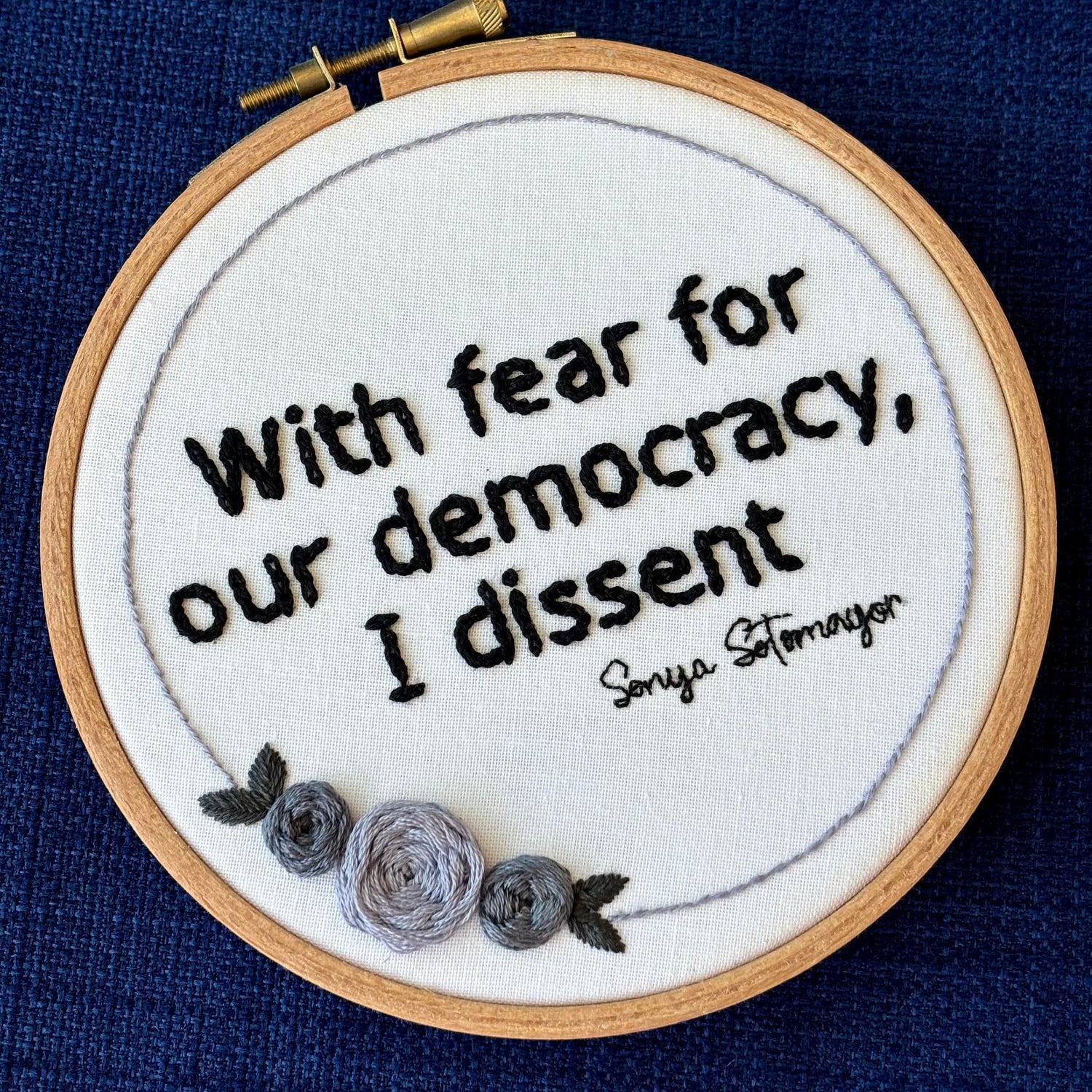 "With fear for our democracy, I dissent. Sonia Sotomayor" Quote hand embroidered in a strong sans-serif font in black embroidery thread. A floral wreath of light and medium gray roses surrounds the text. Fabric is white cotton. Embroidery is displayed as wall art in a wood embroidery hoop.
