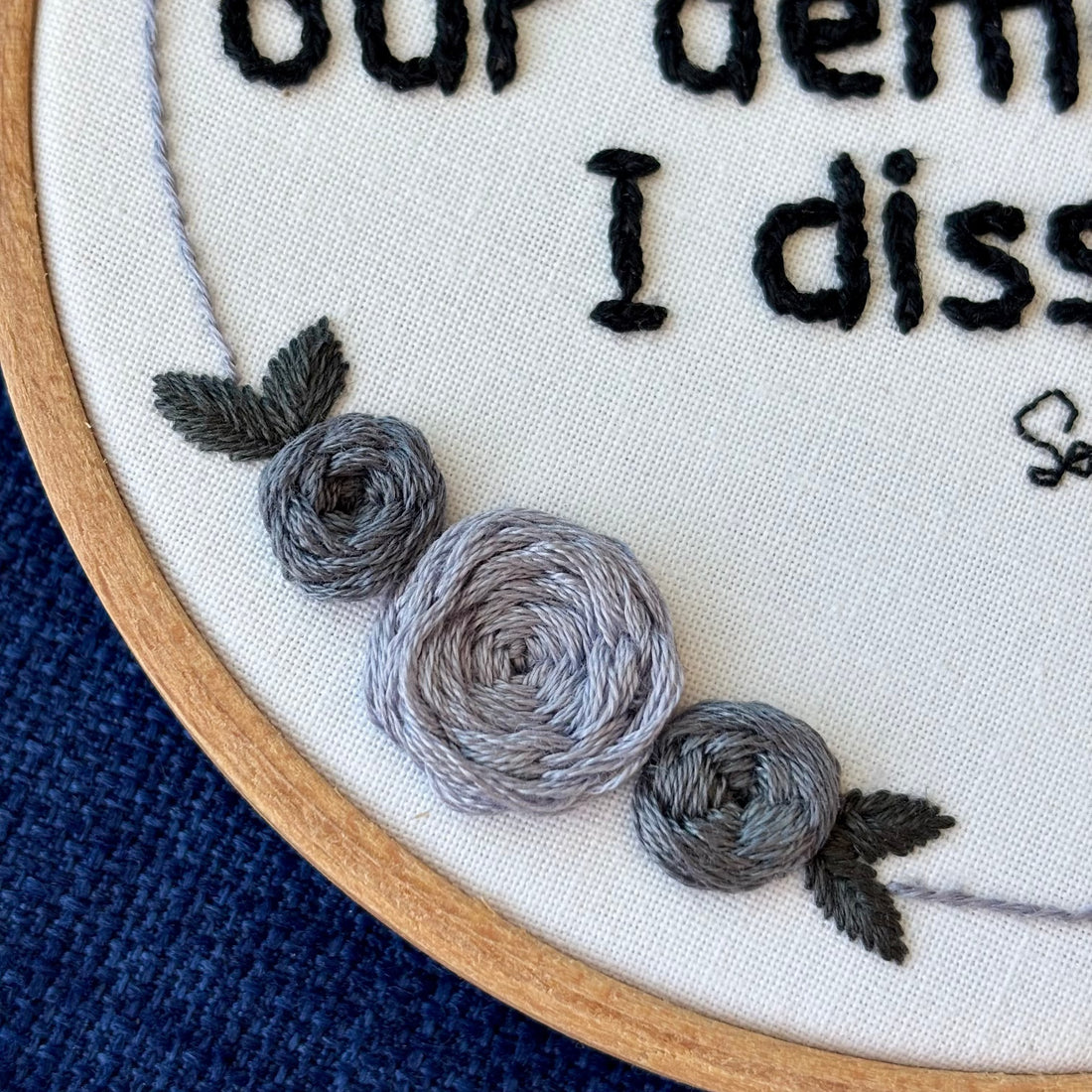 "With fear for our democracy, I dissent. Sonia Sotomayor" Quote hand embroidered in a strong sans-serif font in black embroidery thread. A floral wreath of light and medium gray roses surrounds the text. Fabric is white cotton. Embroidery is displayed as wall art in a wood embroidery hoop.