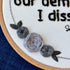 "With fear for our democracy, I dissent. Sonia Sotomayor" Quote hand embroidered in a strong sans-serif font in black embroidery thread. A floral wreath of light and medium gray roses surrounds the text. Fabric is white cotton. Embroidery is displayed as wall art in a wood embroidery hoop.